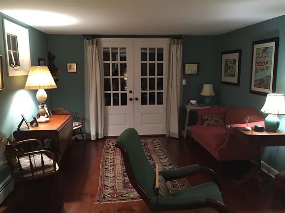 A cozy, teal-walled room with French doors, antique furniture, Persian rug, and soft lighting.