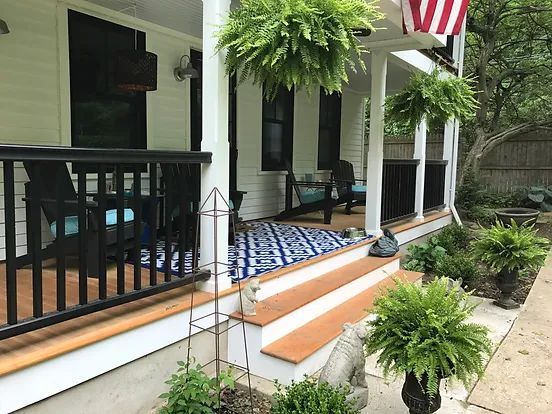 Front porch with ferns, steps, and a blue rug. White siding, black railing, and American flag.