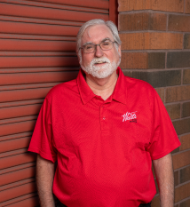 A man with a beard and glasses is wearing a red shirt and standing in front of a brick wall. | Walt's Auto Service
