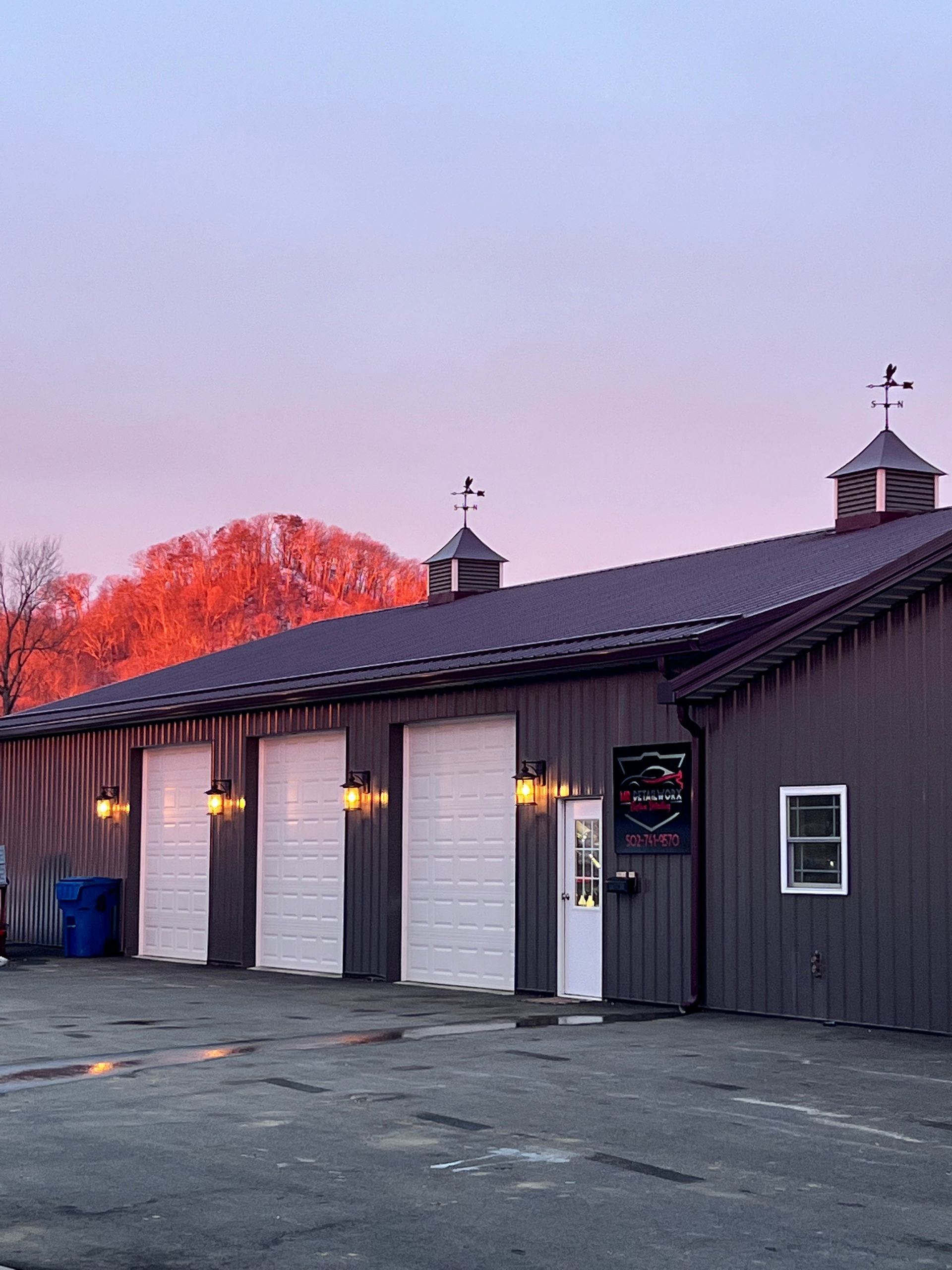 Gray building with white garage doors, a door, and small cupolas, set against a pink and orange sky.