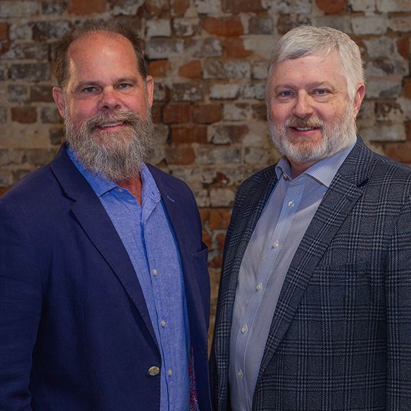 Two men standing together in front of a brick wall, both in suit jackets, smiling.