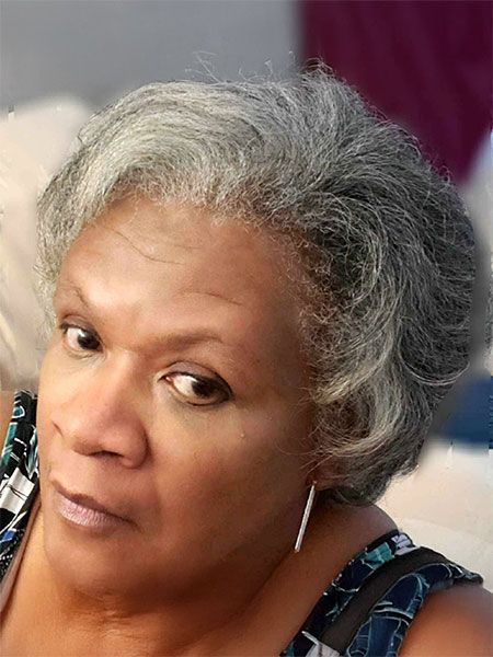 A person with short, grey, textured hair looks toward the side while wearing patterned clothing and silver hoop earrings.