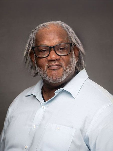 A person with grey locs and glasses wears a light blue button-up shirt against a plain grey background.