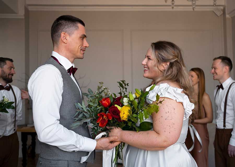 A couple in formal attire holds a colorful bouquet, smiling at each other, flanked by two guests in an indoor setting.