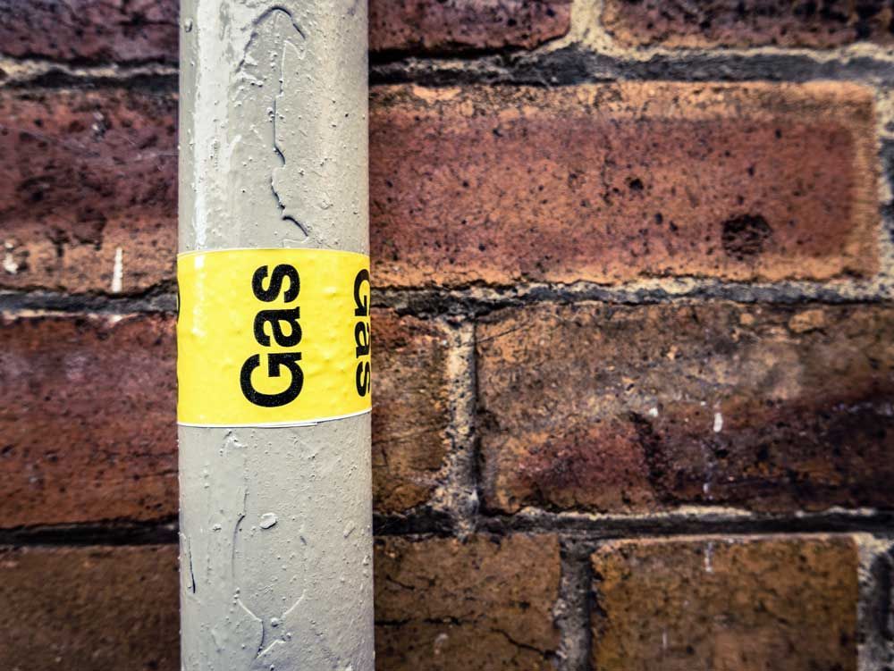 A gas pipe with a yellow sticker on it is against a brick wall.