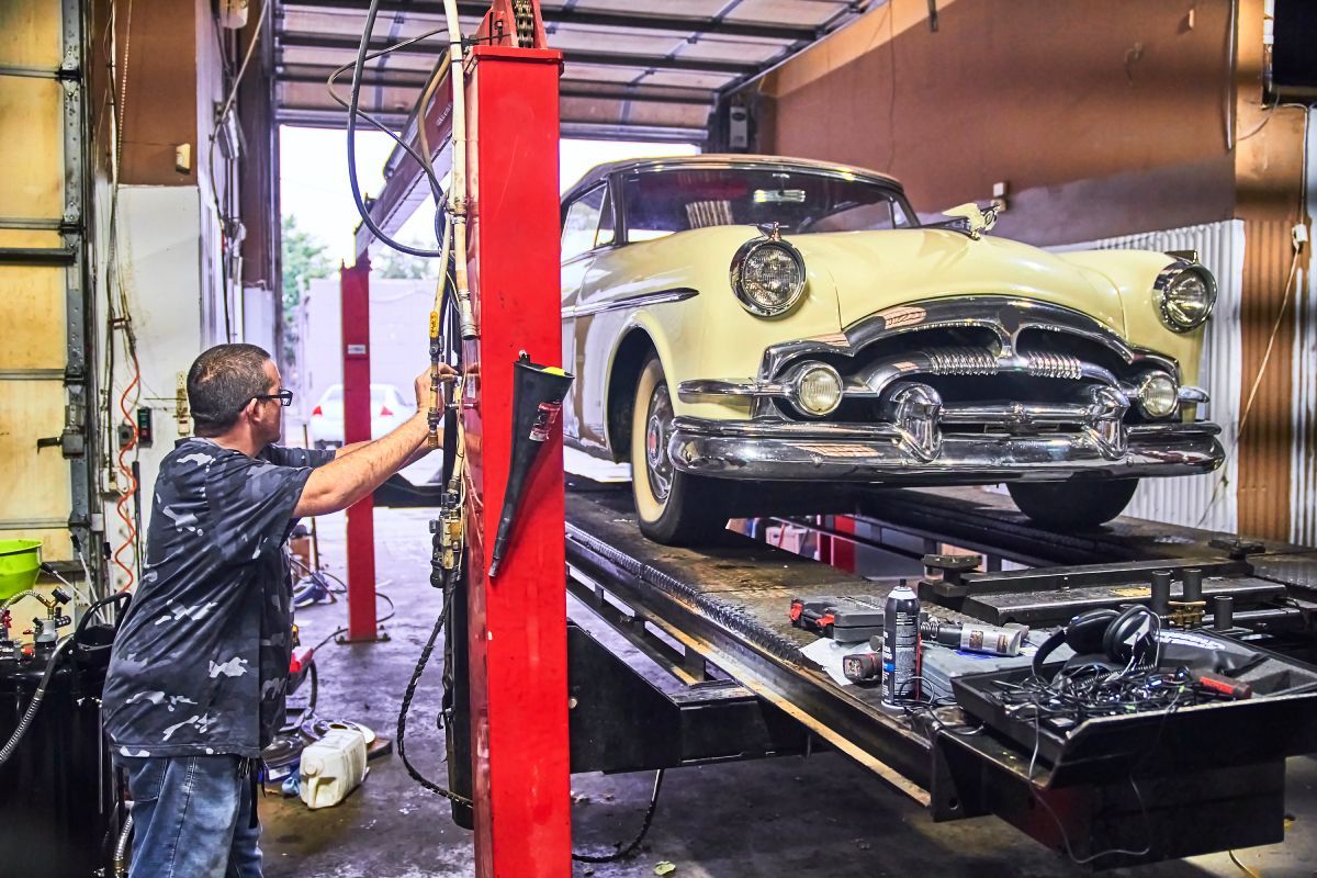 Mechanic working on a classic cream-colored car raised on a lift inside a garage. | Priced Right Auto Clinic