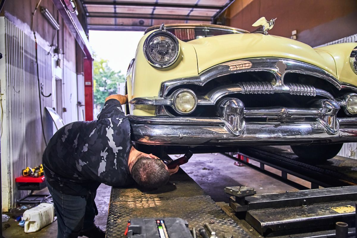 Mechanic working on the underside of a classic yellow car in a garage. | Priced Right Auto Clinic