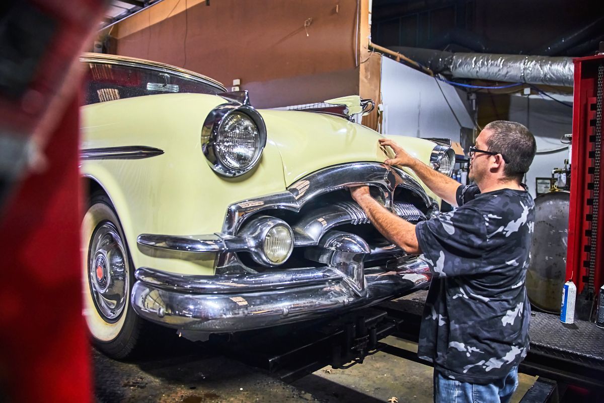 Man working on a light yellow classic car's front bumper, inside a garage. | Priced Right Auto Clinic