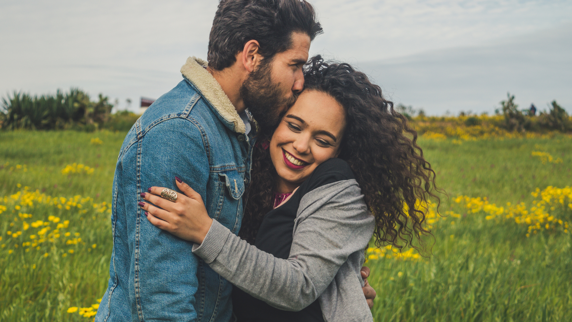 Man kissing a woman's forehead while hugging her in a field of yellow flowers.