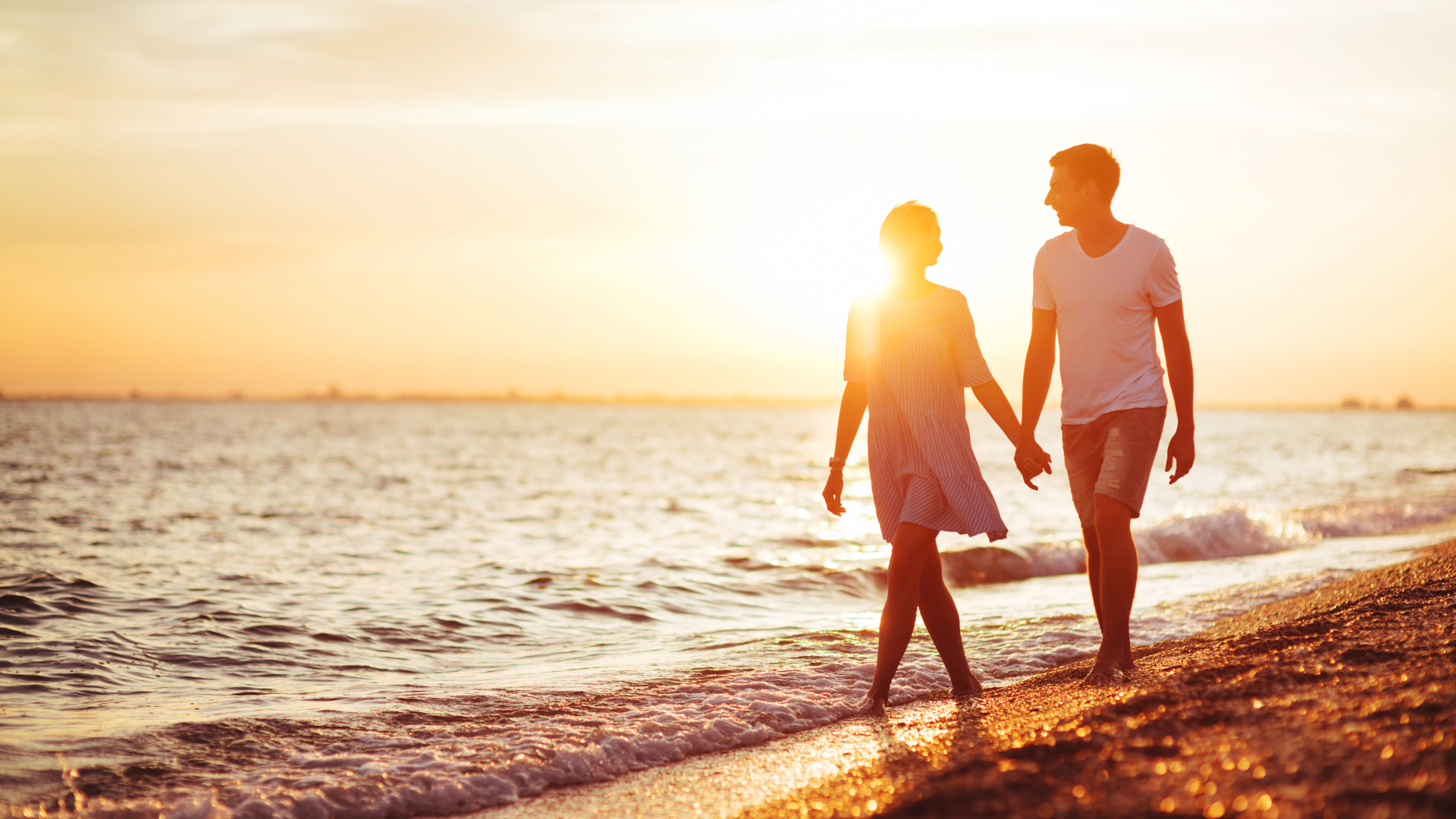 Couple holding hands, walking along a beach at sunset.