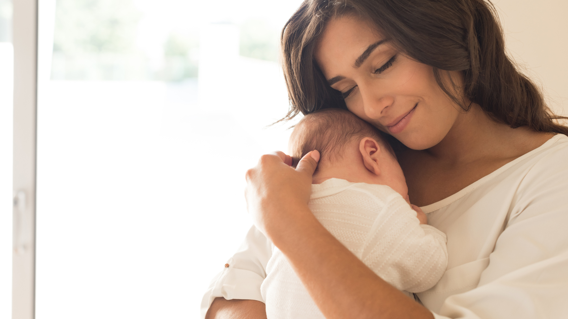 Woman cradles a sleeping baby, softly smiling, near a bright window.
