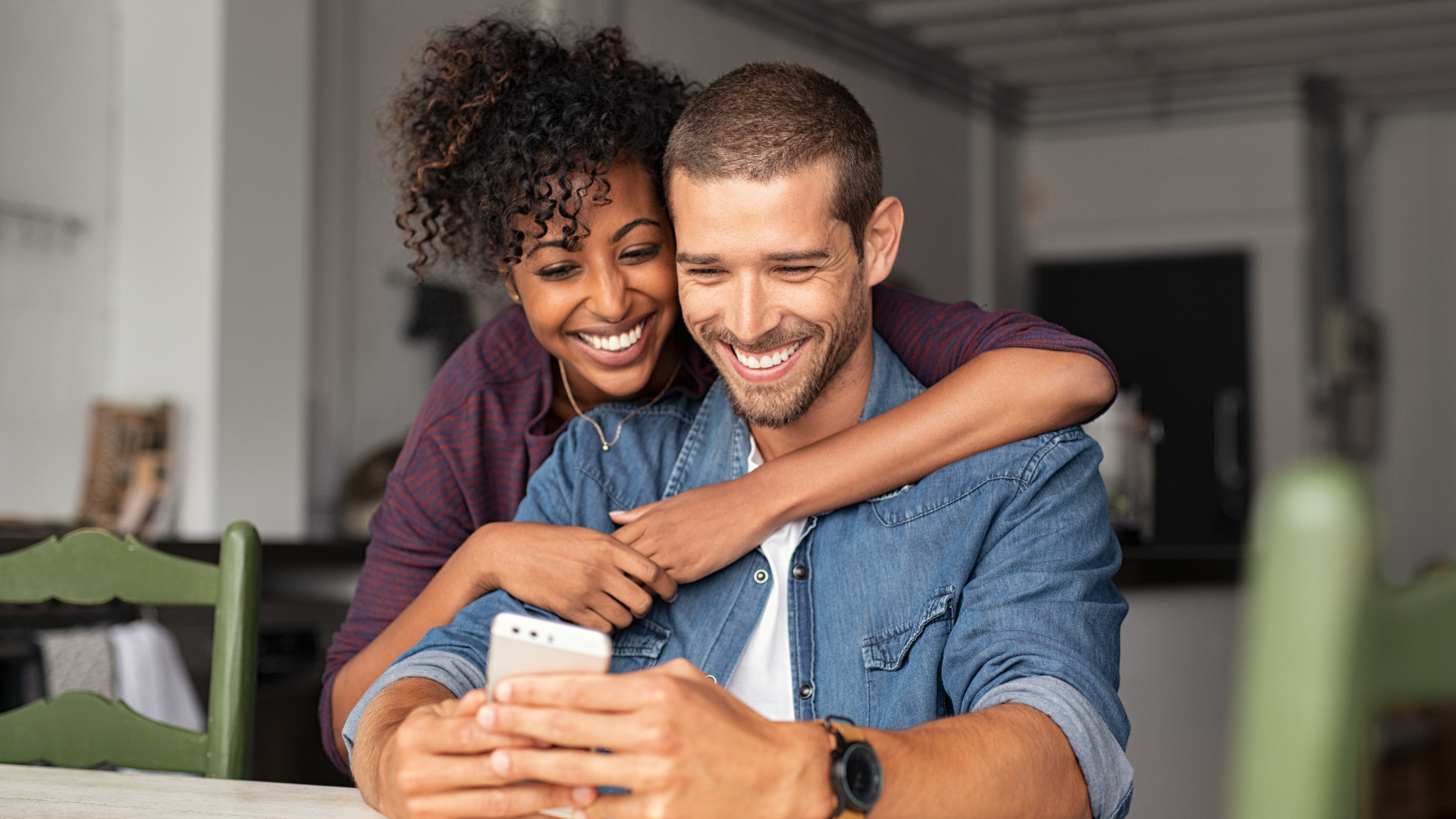 Woman embraces man, both smiling and looking at a smartphone. They are indoors.