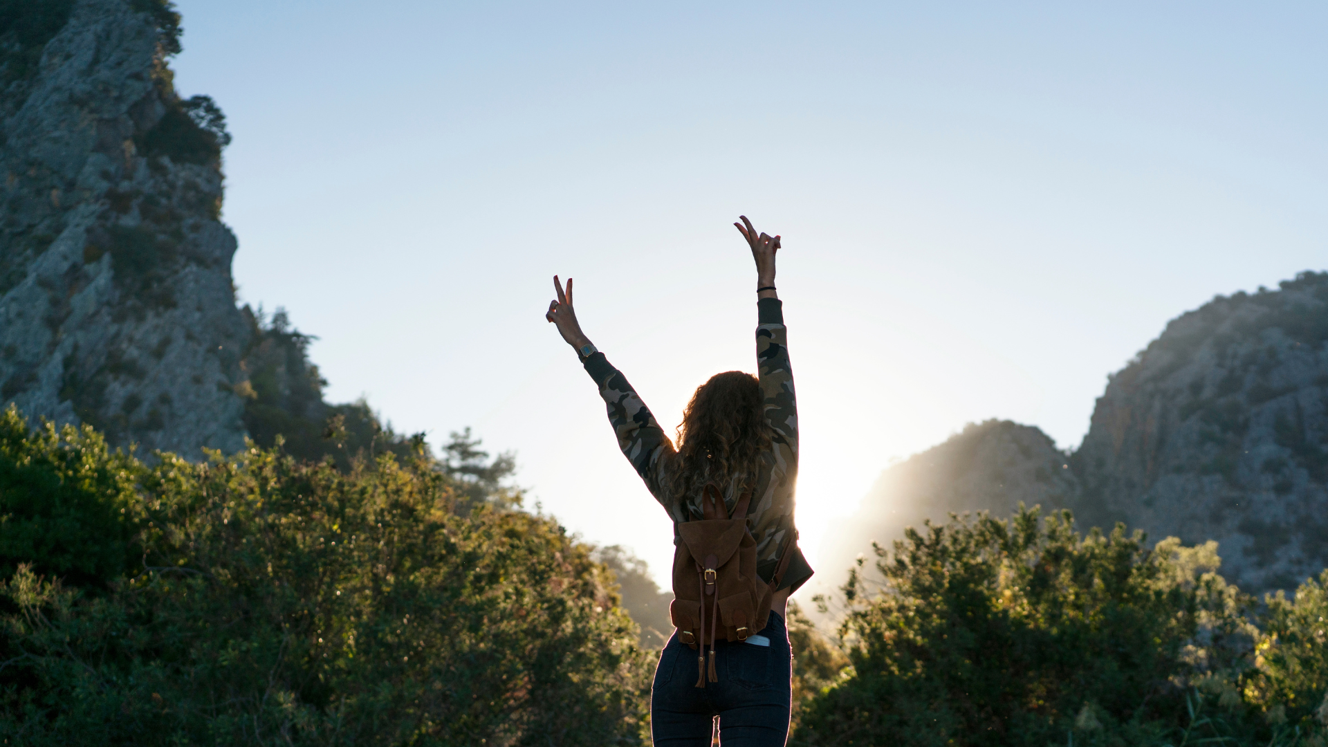 Woman with arms raised making peace signs, standing before a mountain and setting sun.