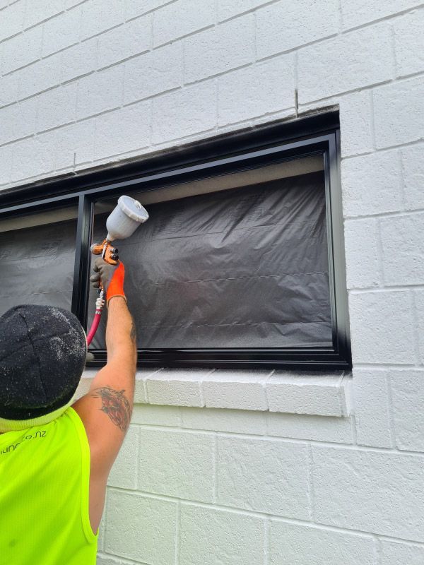 Person spray painting black window frame, covering glass with black plastic, against white brick wall.