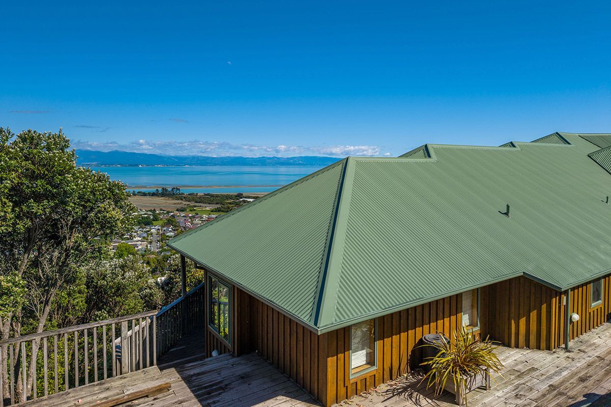 House with green roof and wooden siding overlooking water and mountains on a sunny day.