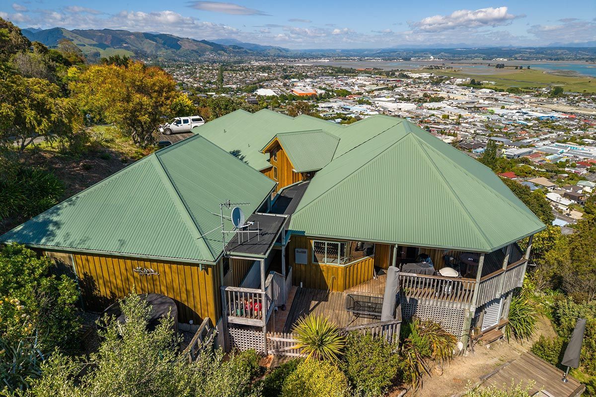 House with green roof sits on a hill overlooking a city and water under a blue sky.