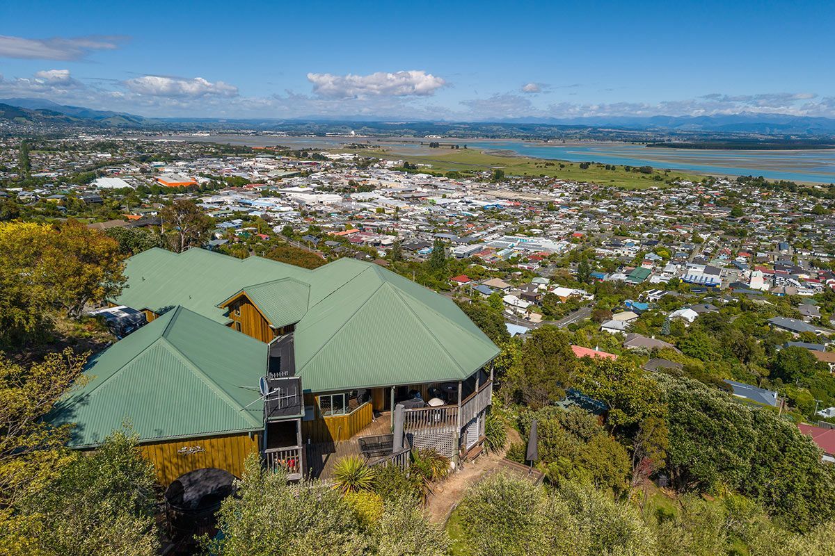White two-story house with black roof and multiple windows, set on a green grassy hill under a cloudy sky.