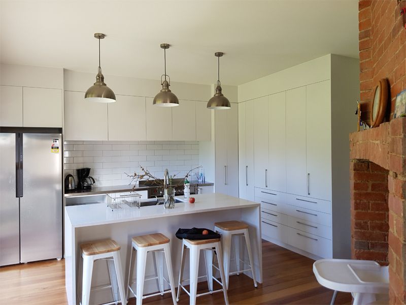 Modern white kitchen with island, stools, stainless steel appliances, pendant lights, and brick fireplace.