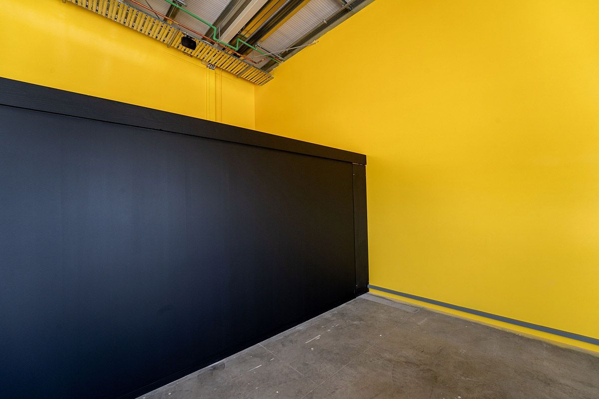 Black wall corner against bright yellow walls, gray floor, industrial ceiling.