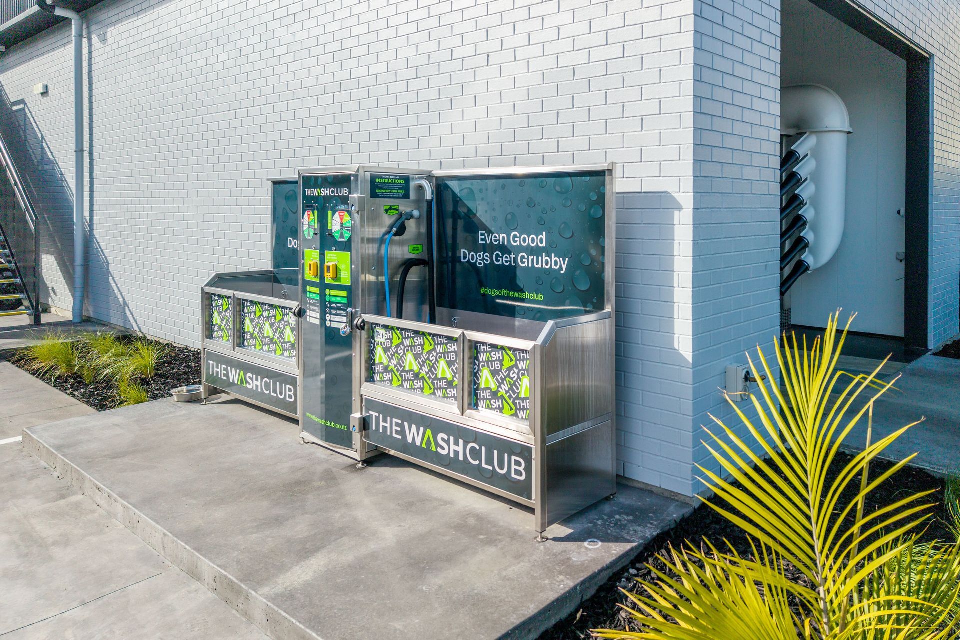 Stainless steel outdoor recycling bins against a white brick wall.