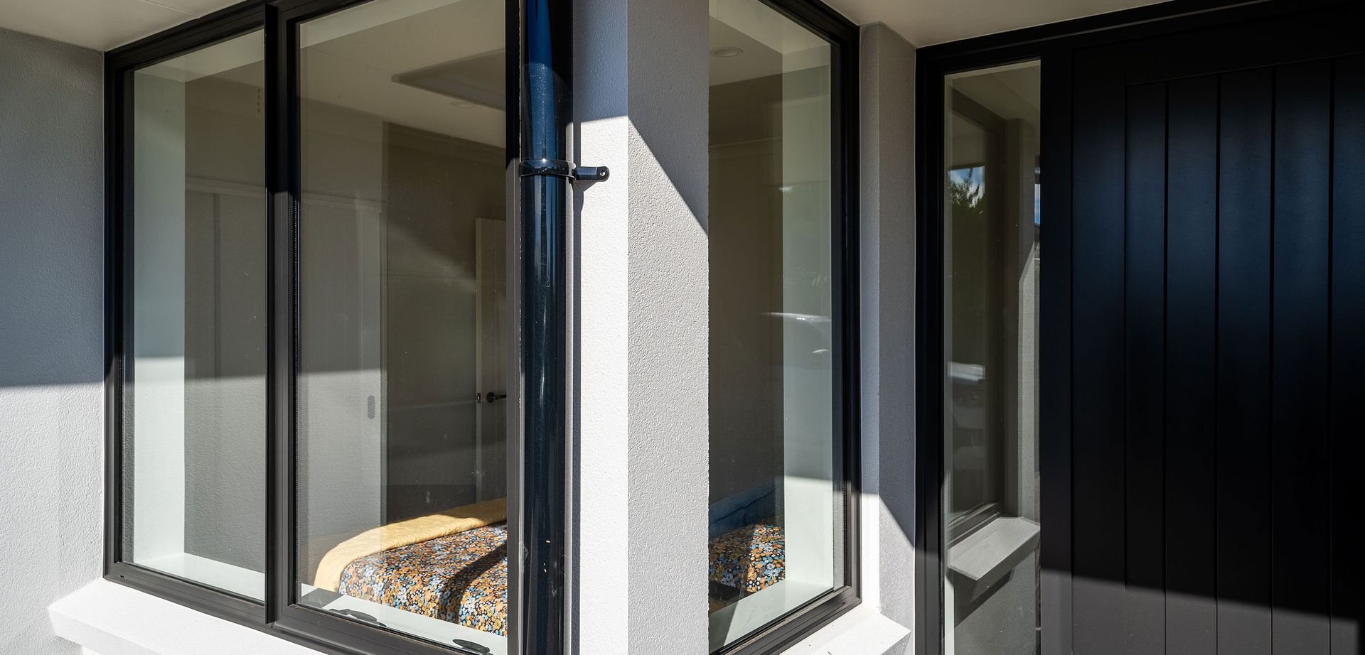 Exterior corner of a building with black-framed windows and a black door; a black downspout runs along the stucco wall.