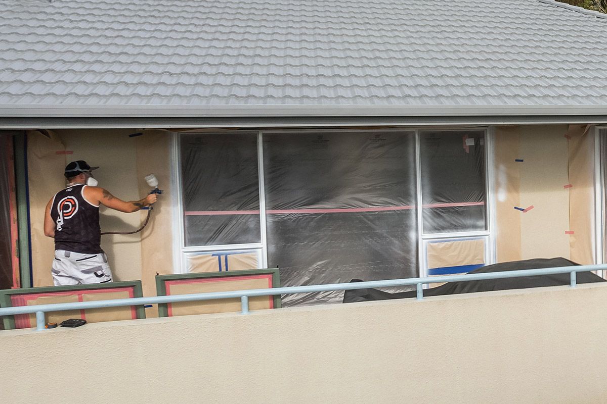 Person sprays exterior wall; window covered in plastic, house roof visible.