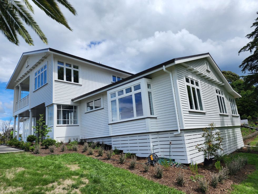 White two-story house with black roof and multiple windows, set on a green grassy hill under a cloudy sky.