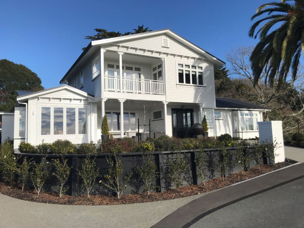 White two-story house with a balcony and black fence, surrounded by greenery.
