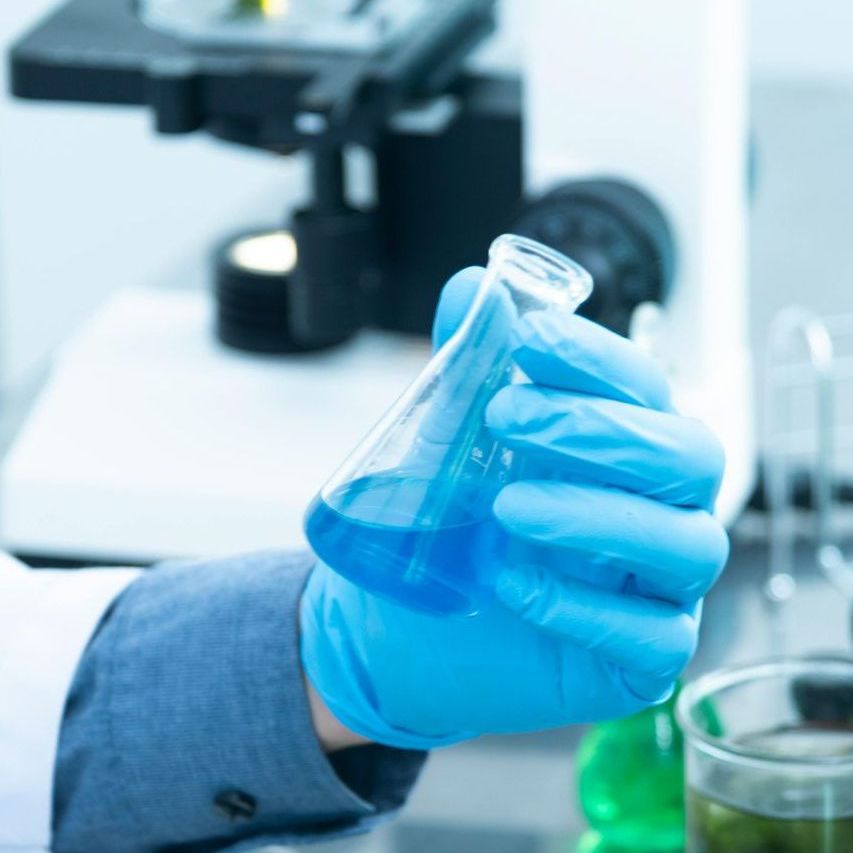 A person wearing blue gloves is holding a beaker of blue liquid in front of a microscope