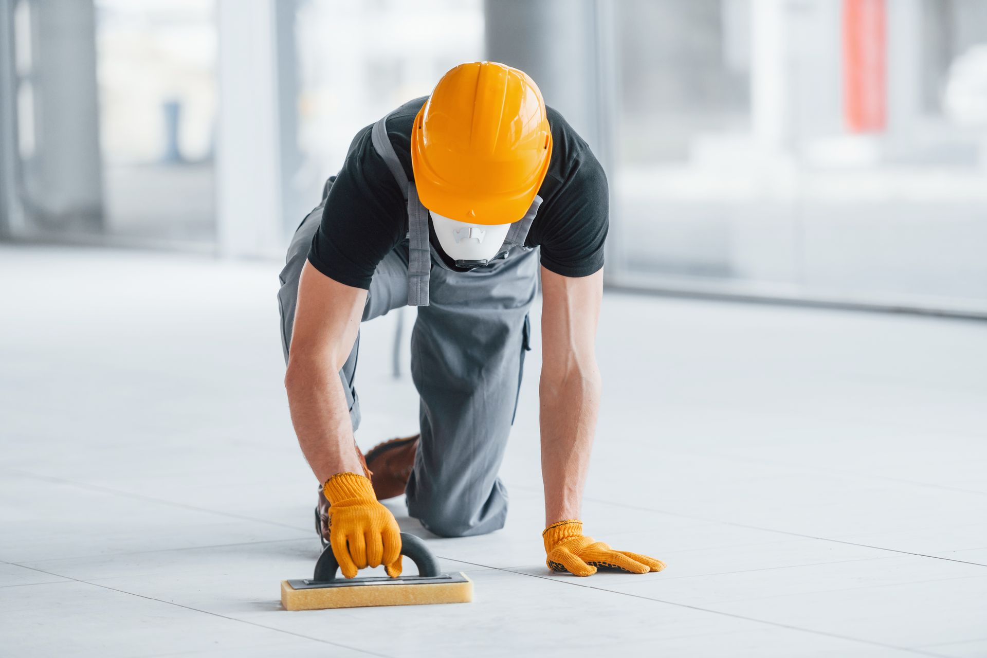 A construction worker is kneeling on the floor while using a trowel.