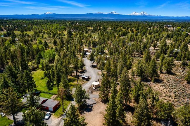 Aerial view of a forested campsite with cabins, dirt roads, and distant snowcapped mountains under a clear blue sky