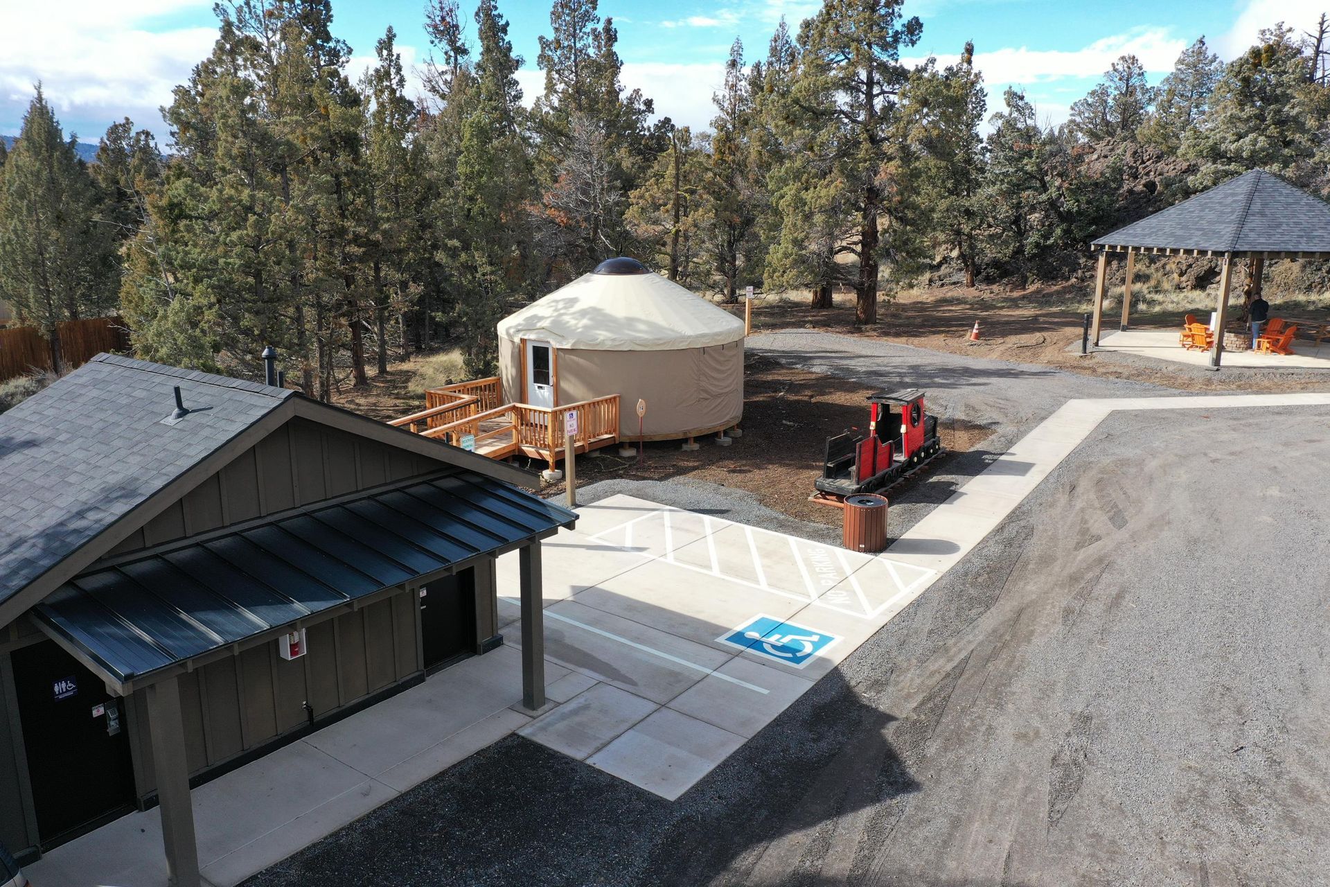 Outdoor campsite with a yurt, pavilion, picnic area, and paved parking lot surrounded by trees