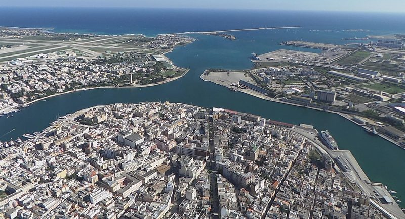 Aerial view of a coastal city with waterways, harbor, buildings, and the sea under a blue sky.
