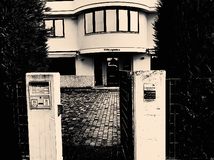 Yellow building with black windows, red brick path, gated entrance, and trees.
