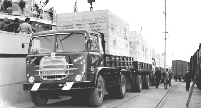 Truck loaded with crates at a dock. Workers by the truck and alongside a ship. Railroad cars in the distance.