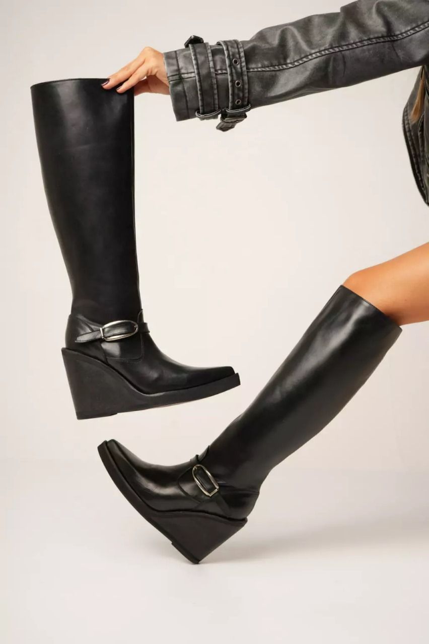 Una mujer lleva botas de cuña de cuero negro. Una bota se sostiene con una mano; la otra, sobre la pierna.