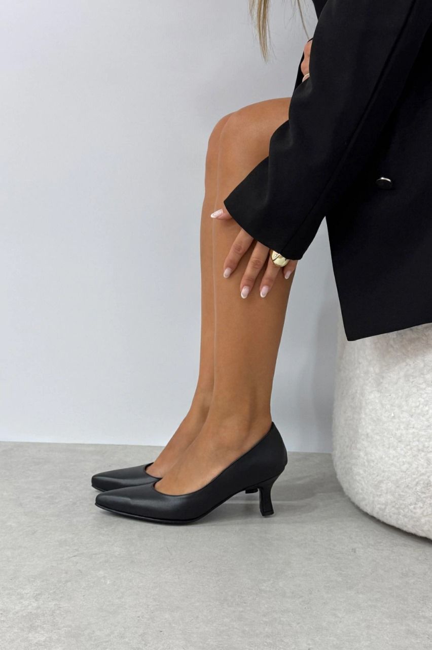 Piernas de mujer, con tacones negros, sentada cerca de una otomana blanca y un blazer negro.