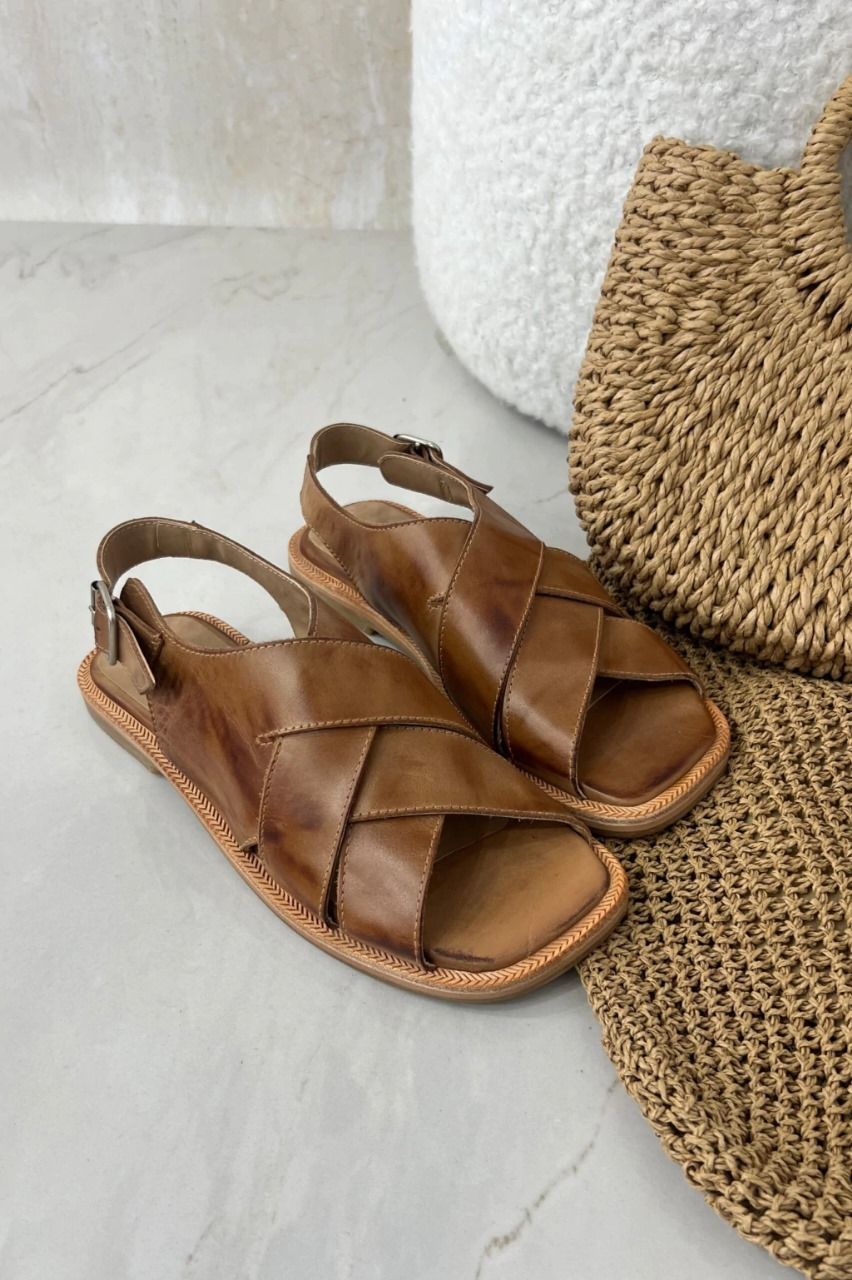 Sandalias de piel marrón con tiras cruzadas, junto a un bolso tejido y fondo blanco texturizado.