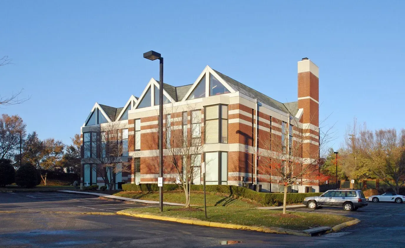 Brick academic building with white peaked roof trusses and a red tower beside a parking lot in autumn.