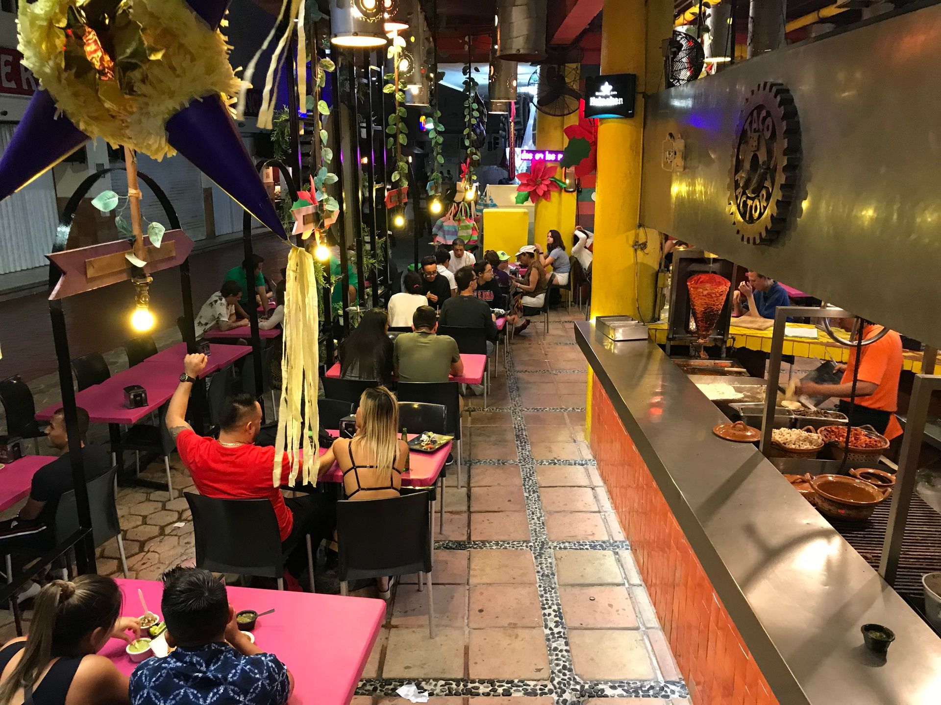 Restaurante con mesas rosas y clientes cenando. Barra con cocinero. Iluminación superior y decoración. taco factory