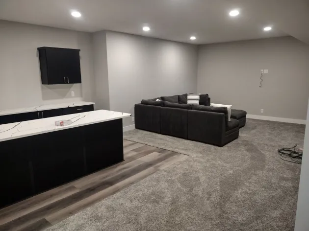 Modern basement entertainment space: dark sectional sofa, black cabinets, white countertop, gray walls, carpet.