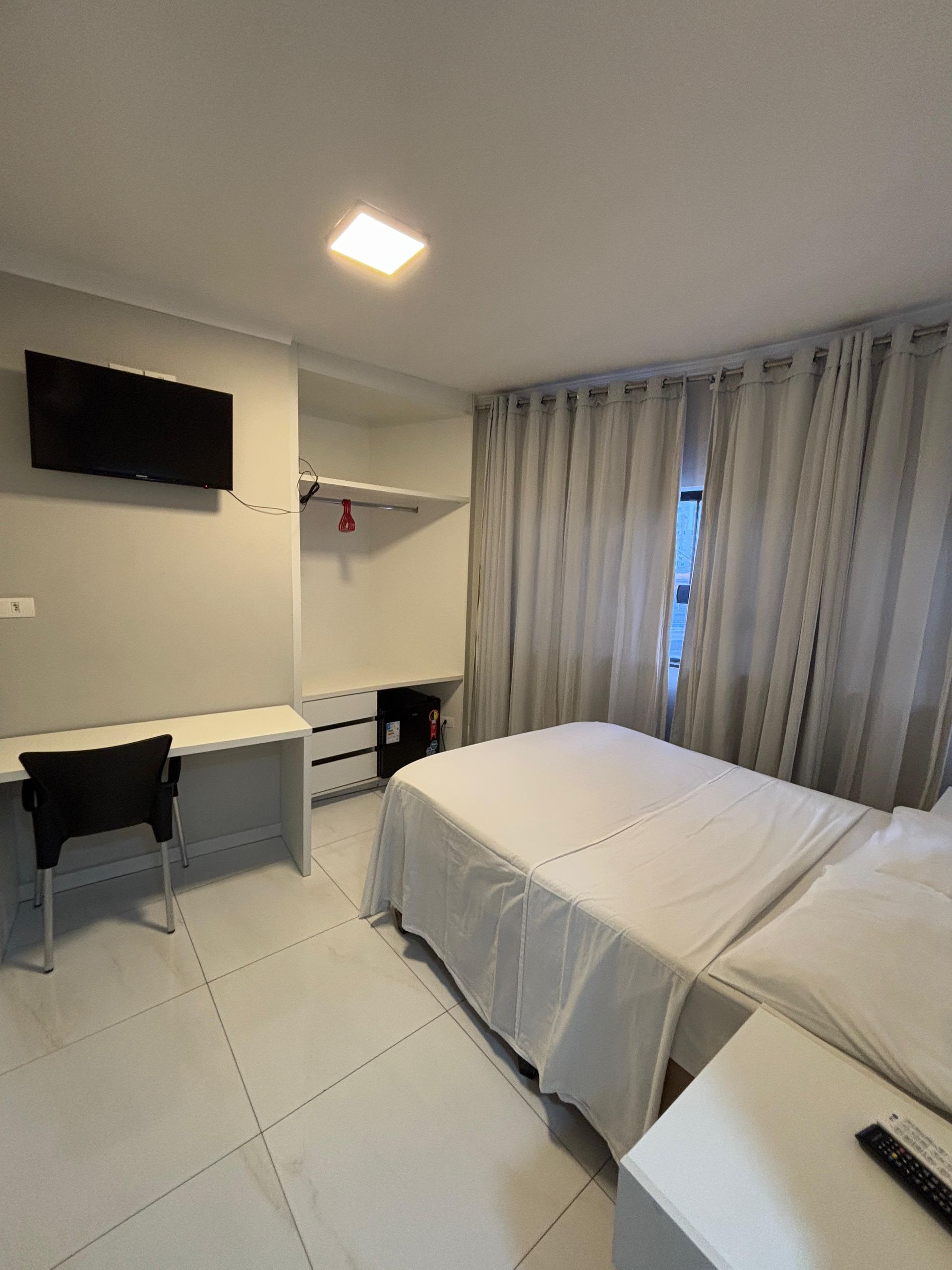 Quarto com cama branca, escrivaninha, TV, armário e janela.