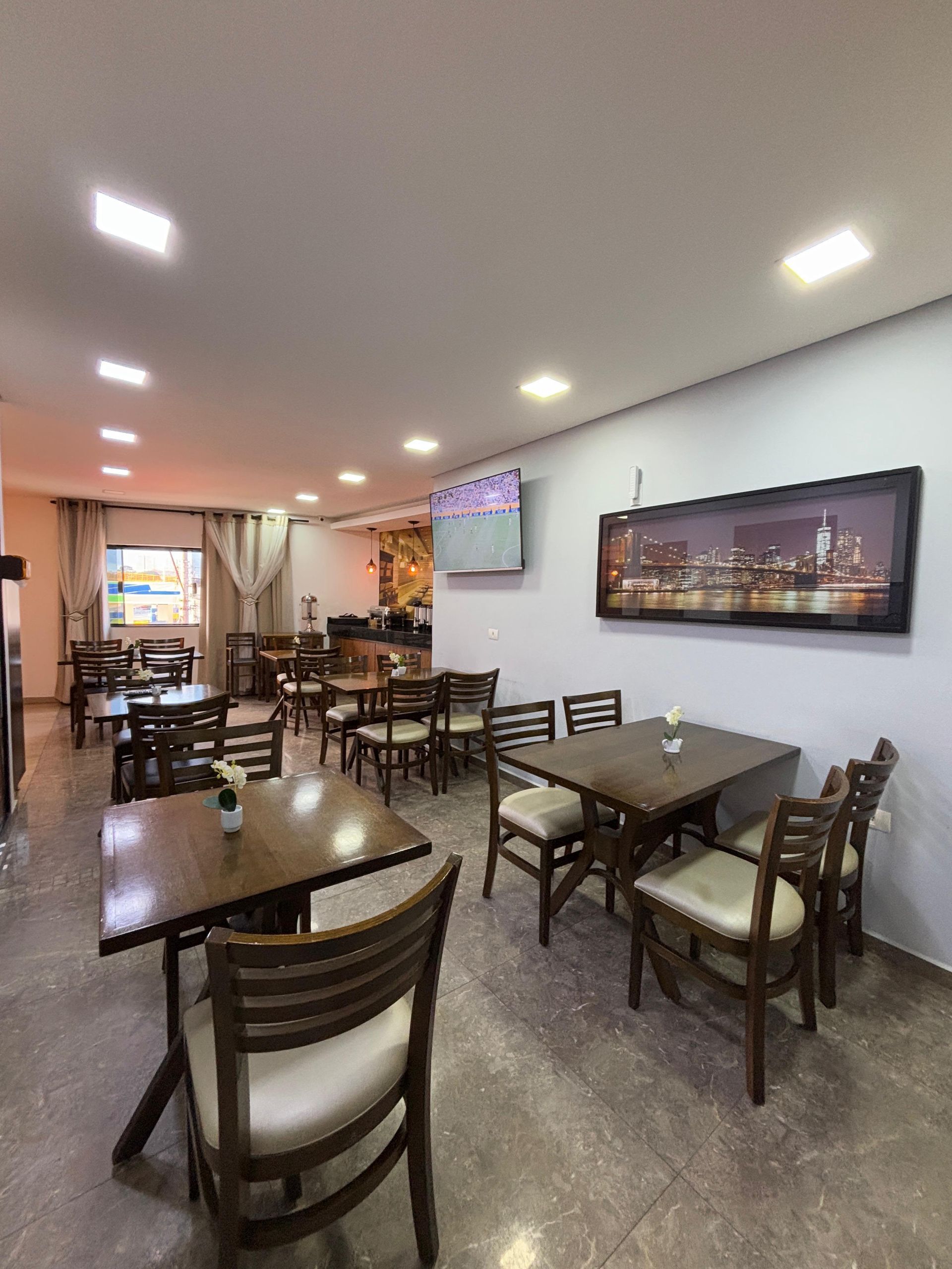 Interior de restaurante com mesas e cadeiras de madeira, uma televisão e obras de arte com paisagens urbanas.
