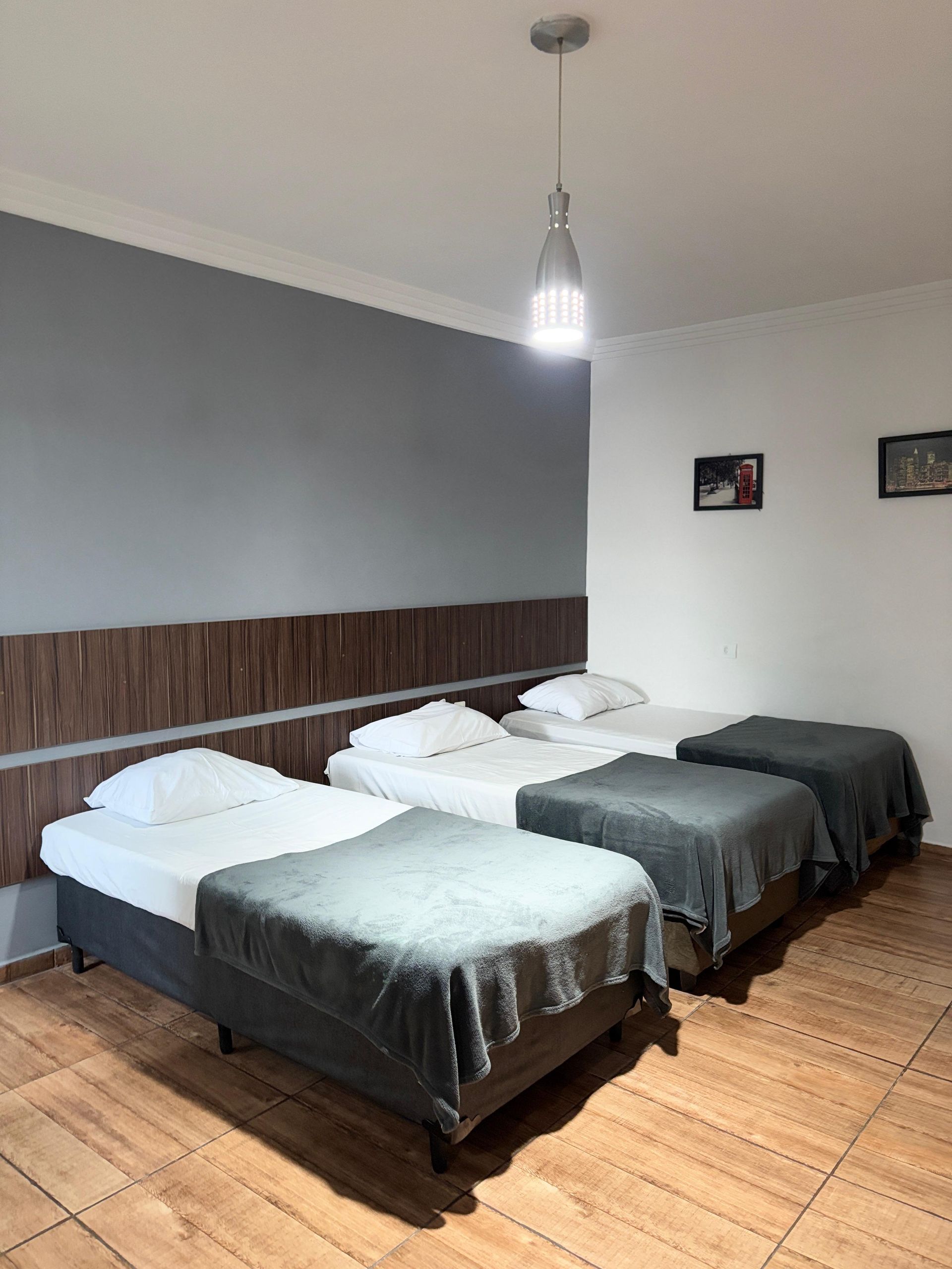 Três camas em um quarto de hotel com cobertores cinza, cabeceira de madeira e luminária.