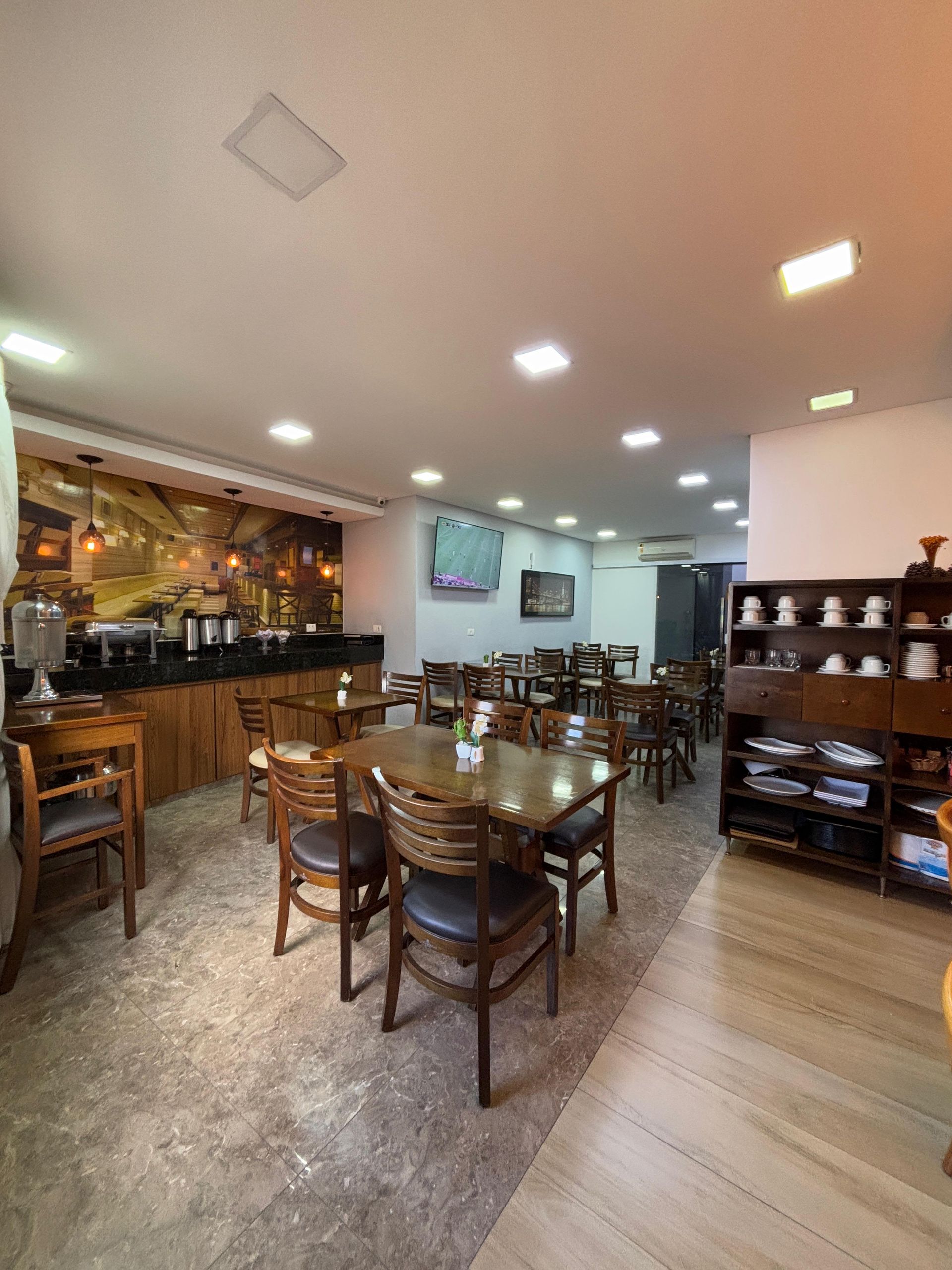 Área de jantar do restaurante com mesas, cadeiras e buffet. Mobiliário marrom, paredes em tons neutros, TV.