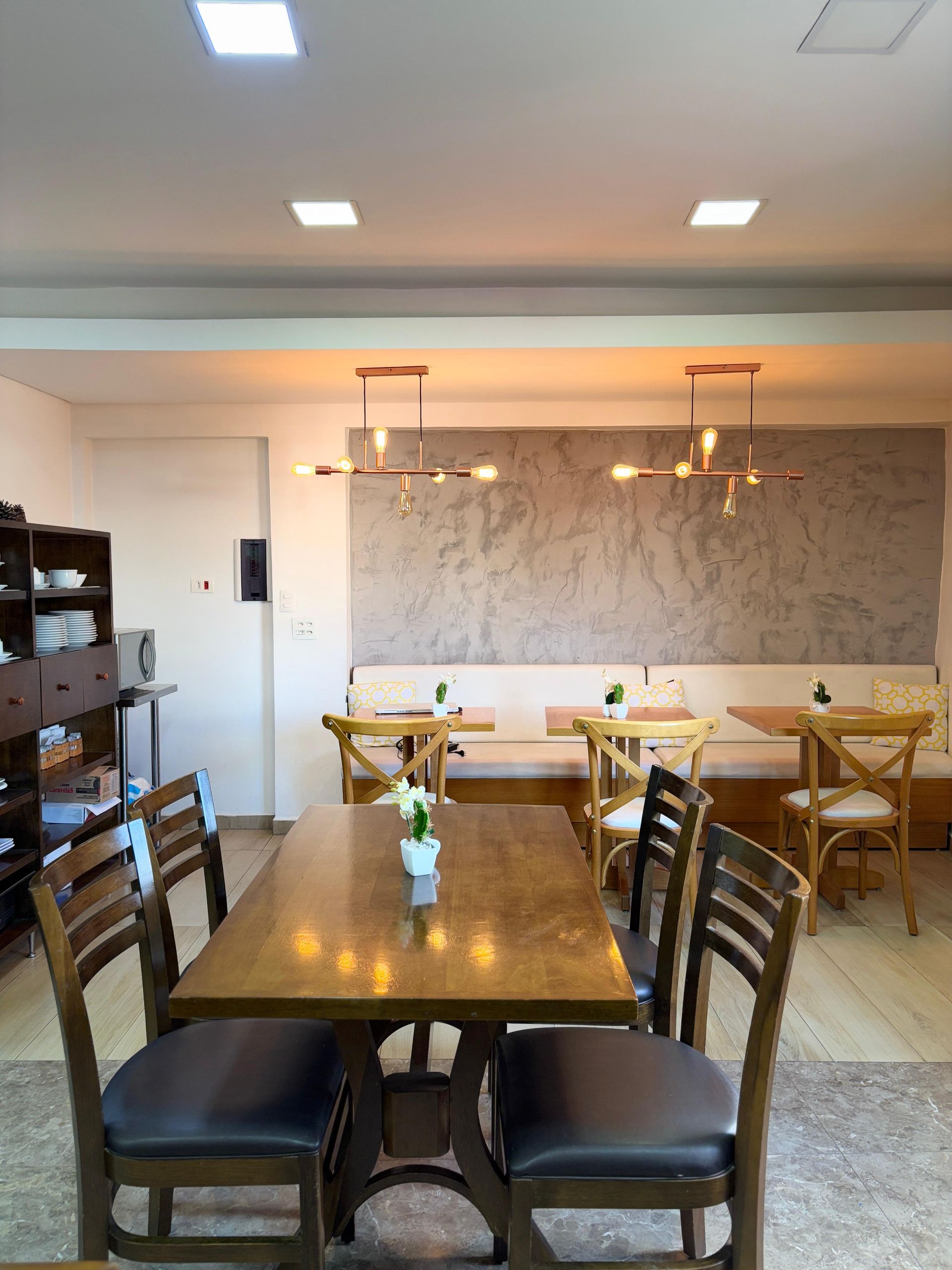 Interior de restaurante com mesas, cadeiras e bancos de madeira, iluminado por luzes no teto.