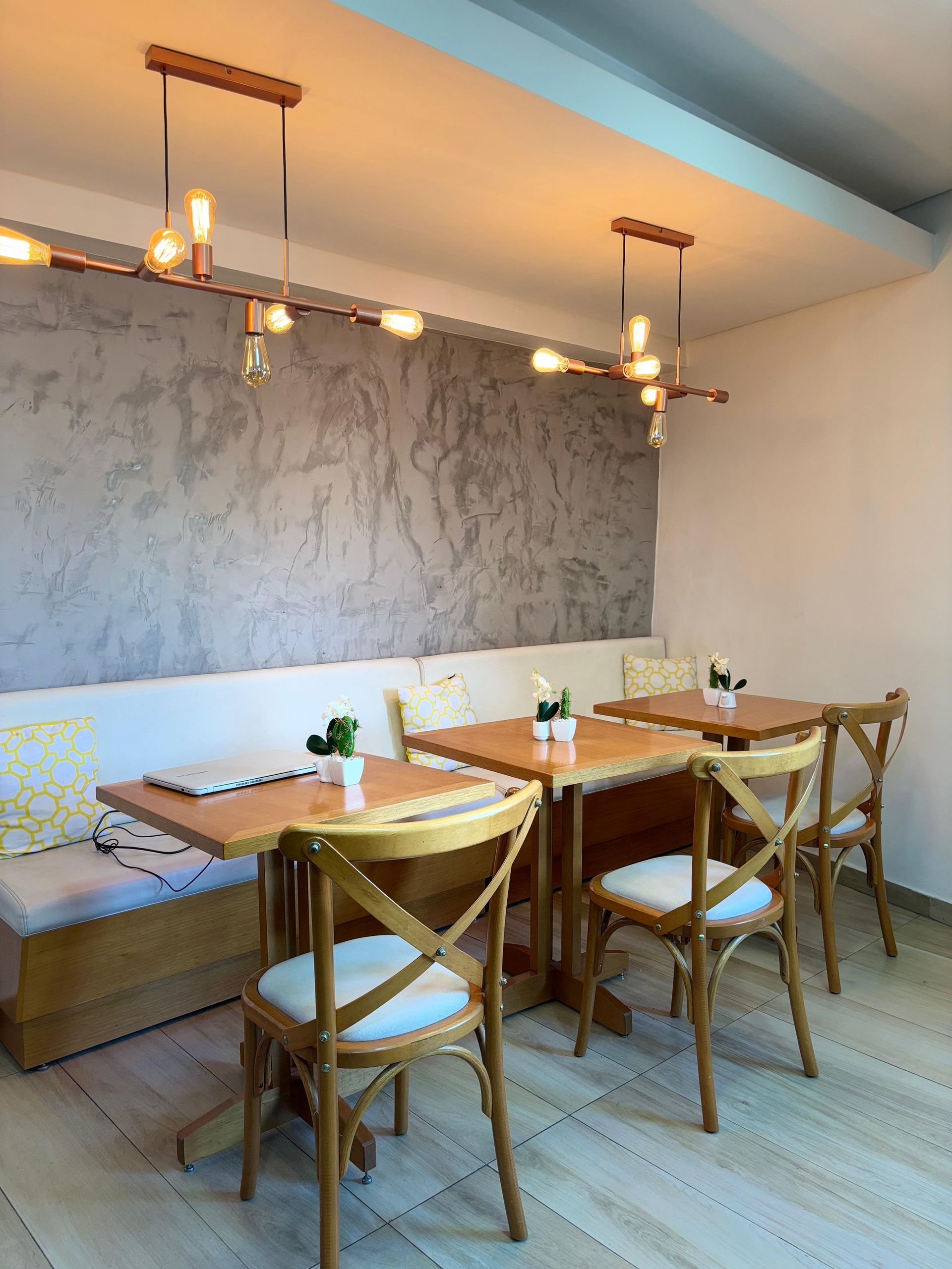 Interior de café com mesas e cadeiras de madeira, um banco embutido e iluminação decorativa contra uma parede cinza texturizada.