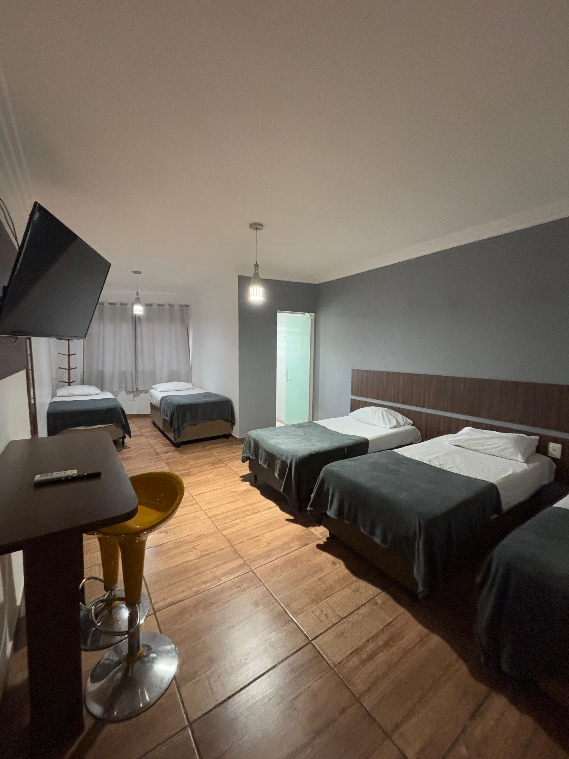 Quarto de hotel com várias camas, cobertores cinza-escuros, piso de madeira e um banco de bar amarelo.