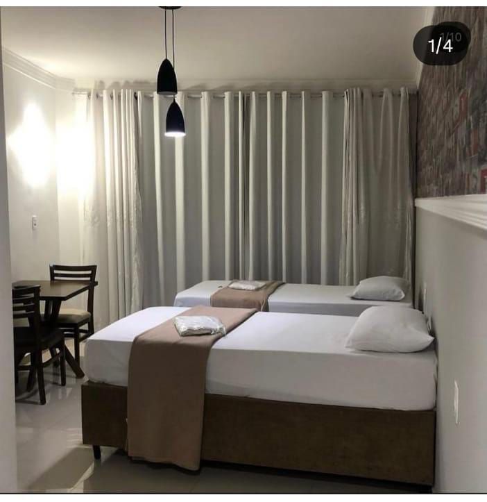 Um pequeno quarto com duas camas, cortinas brancas e uma pequena mesa com cadeiras.