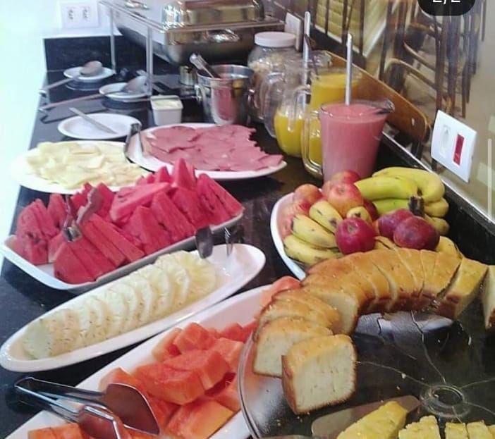 Buffet de café da manhã com frutas, pão, presunto, suco e ovos em uma bancada preta.